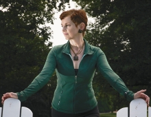 Catherine Waters Raymond, a woman with red hair wearing a green shirt, stands between two white chairs.